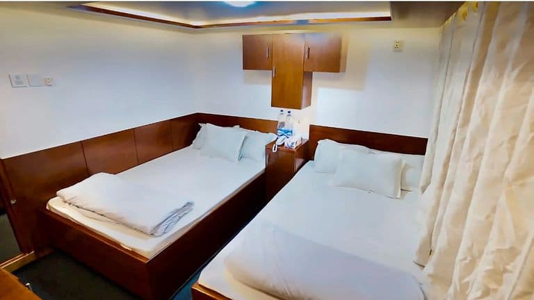 A cabin with two beds side by side, white pillows and cream curtains by the window