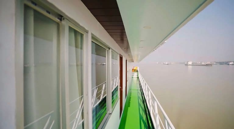 The open side deck of the vessel in daylight, with a green floor, a white railing and the river beyond